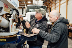 Two workers performing maintenance on industrial equipment in a workshop with a truck visible in the background.