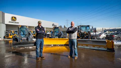 Two men standing in front of yellow snow plows at a John Deere equipment dealership lot