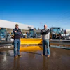 Two men standing in front of yellow snow plows at a John Deere equipment dealership lot