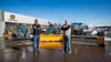 Two men standing in front of yellow snow plows at a John Deere equipment dealership lot
