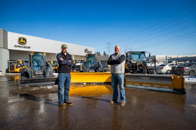 Two men standing in front of yellow snow plows at a John Deere equipment dealership lot