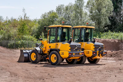 Yellow Volvo wheel loader and vibratory rollers parked on construction site with trees in background