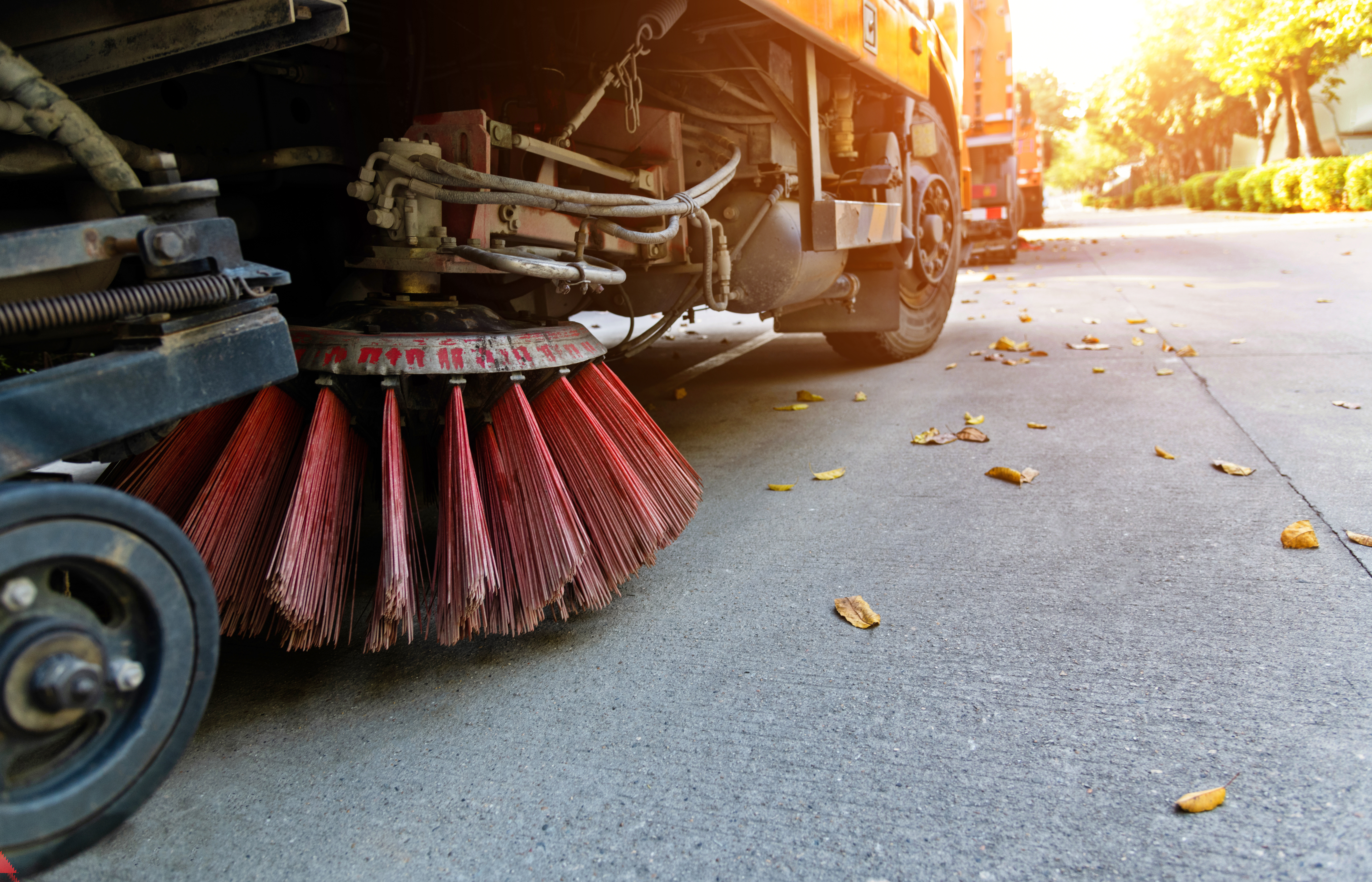 Street sweeper truck with red rotating brush cleaning pavement on sunny day